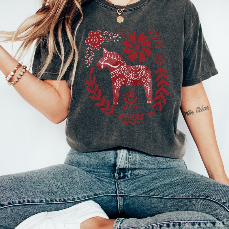 May include: A dark grey t-shirt featuring a red and white Scandinavian Dala horse design. The horse is surrounded by red floral designs.