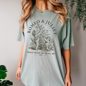 May include: A light blue t-shirt with a black graphic print of two cherubs holding a flower branch. The text "Romeo & Juliet" is printed in a circle around the cherubs. The text "What must be shall be" is printed below the cherubs.