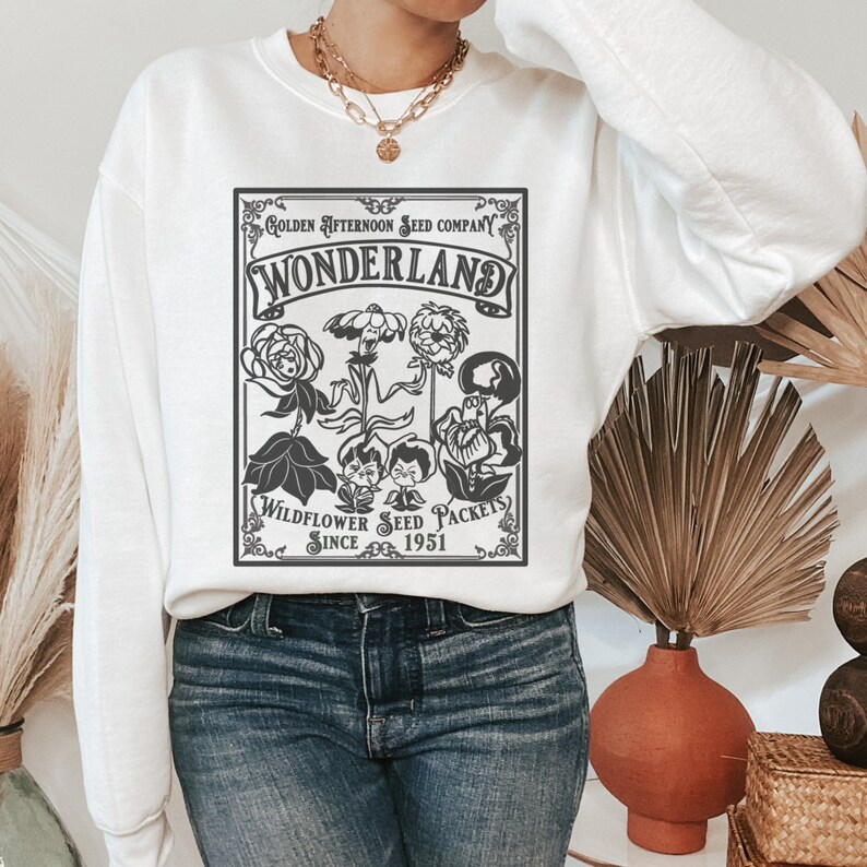May include: White sweatshirt with a black and white graphic print of a vintage seed packet. The packet says "Golden Afternoon Seed Company Wonderland Wildflower Seed Packets Since 1951".