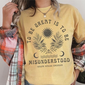 Op de afbeelding: Geel T-shirt met een vintage-stijl grafische afbeelding met de tekst "To be great is to be misunderstood - Ralph Waldo Emerson". De afbeelding bevat een zon, twee palmbladeren en twee halve manen.