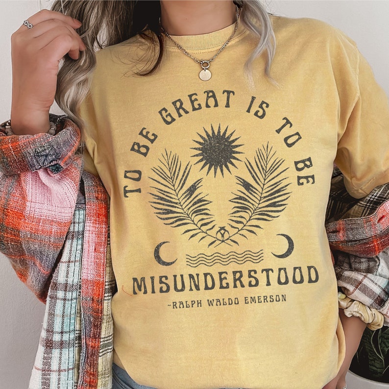 Op de afbeelding: Geel T-shirt met een vintage-stijl grafische afbeelding met de tekst "To be great is to be misunderstood - Ralph Waldo Emerson". De afbeelding bevat een zon, twee palmbladeren en twee halve manen.