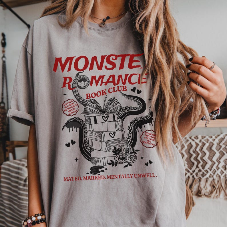 Pu&ograve; includere: T-shirt grigio chiaro con la scritta rossa "MONSTER ROMANCE BOOK CLUB". La grafica presenta una pila di libri, cuori, tentacoli e altro testo. Le maniche della maglietta sono arrotolate.