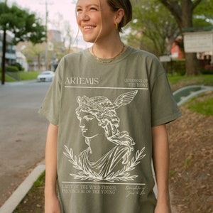 May include: A green t-shirt with a white graphic of Artemis, the Greek goddess of the hunt. The graphic includes a portrait of Artemis with a crown and a bow and arrow. The text "ARTEMIS, GODDESS OF THE HUNT, LADY OF THE WILD THINGS, PROTECTOR OF THE YOUNG, Daughter of Zeus & Leto" is printed on the shirt.