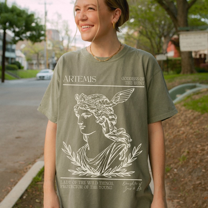 May include: A green t-shirt with a white graphic of Artemis, the Greek goddess of the hunt. The graphic includes a portrait of Artemis with a crown and a bow and arrow. The text "ARTEMIS, GODDESS OF THE HUNT, LADY OF THE WILD THINGS, PROTECTOR OF THE YOUNG, Daughter of Zeus & Leto" is printed on the shirt.