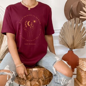 May include: Burgundy t-shirt with a graphic of hands holding a moon and sun with the quote "I took my power in my hand and went against the flow" by Emily Dickinson.