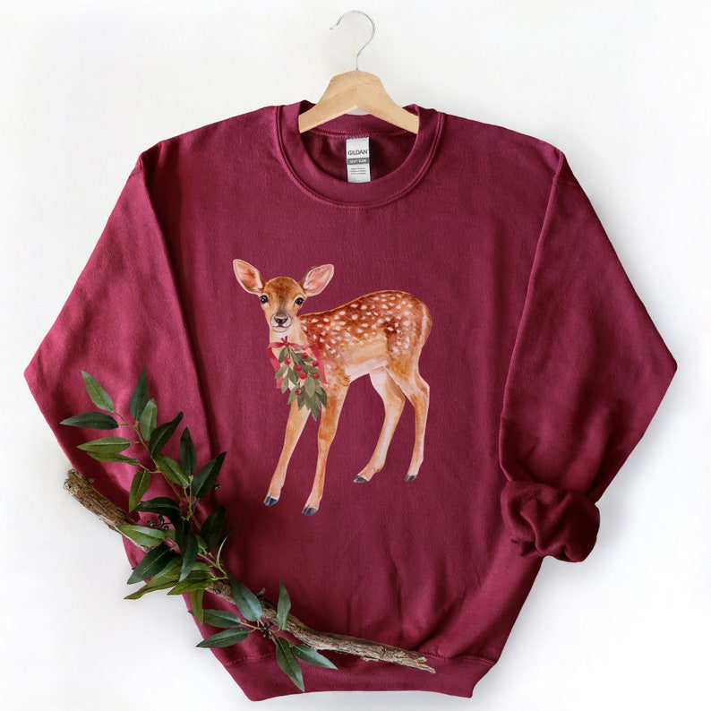 May include: A maroon sweatshirt with a watercolor illustration of a fawn wearing a holly berry garland. The fawn is standing on a white background.