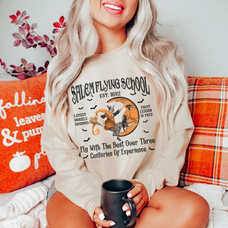 Op de afbeelding: Een beige sweatshirt met een grafische print van een heks die op een bezem voor een halve maan vliegt. De tekst luidt "Salem Flying School Est. 1892" en "Fly With The Best Over Three Centuries Of Experience".