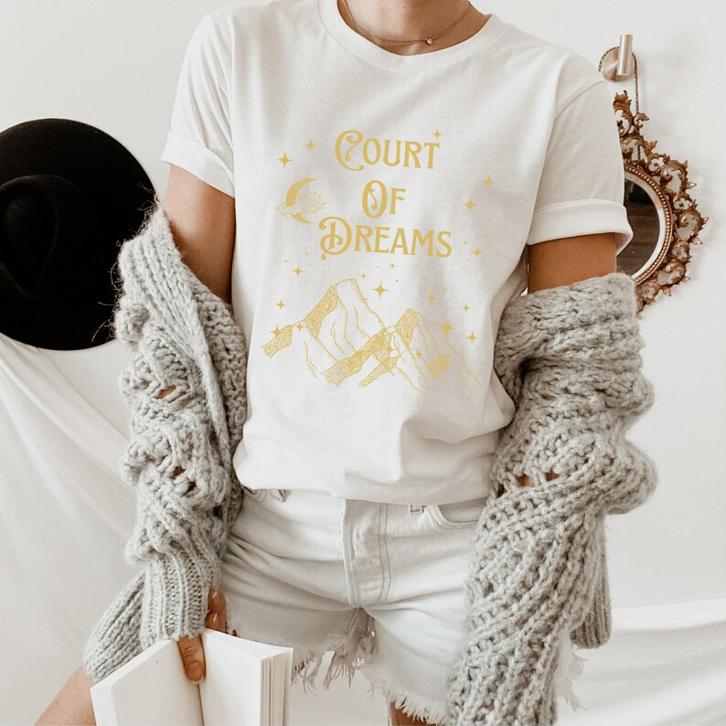 May include: White t-shirt featuring a gold graphic design of mountains, a crescent moon, and stars. The text "COURT OF DREAMS" is printed in gold on the shirt.