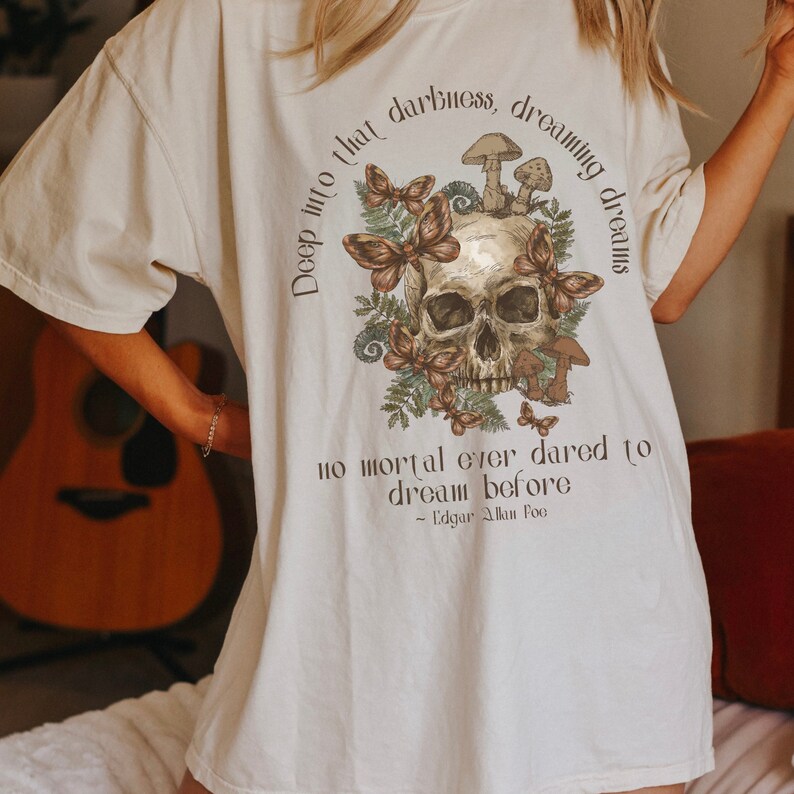 May include: A white t-shirt featuring a skull graphic surrounded by butterflies and mushrooms. The text "Deep into that darkness, dreaming dreams no mortal ever dared to dream before - Edgar Allan Poe" is printed on the shirt.