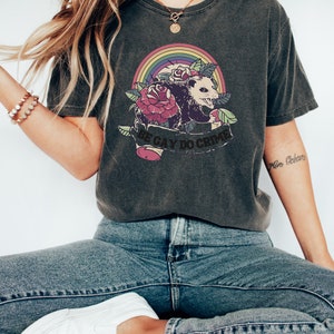 May include: Dark gray t-shirt featuring a graphic of an opossum, roses, and a rainbow. The text "BE GAY DO CRIME" is printed on a banner below the opossum. The shirt is paired with blue jeans and white sneakers.