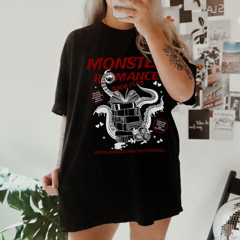 Pu&ograve; includere: T-shirt nero con un design grafico rosso. Il design presenta il testo "MONSTER ROMANCE BOOK CLUB", una pila di libri, tentacoli e cuori. Il testo "MATED. MARKED. MENTALLY UNWELL." &egrave; in basso.