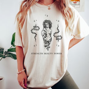 May include: A white t-shirt with a black and white illustration of a woman with snakes and the text "STRENGTH BEAUTY POWER".