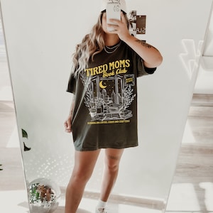May include: A dark green t-shirt with a graphic design that says "Tired Moms Book Club" with a cityscape and a crescent moon. The text "Running on coffee, chaos and chapters" is printed below the graphic.
