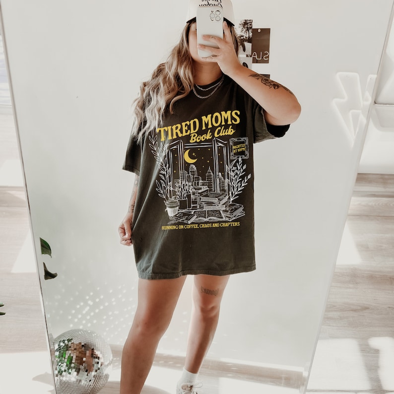 May include: A dark green t-shirt with a graphic design that says "Tired Moms Book Club" with a cityscape and a crescent moon. The text "Running on coffee, chaos and chapters" is printed below the graphic.