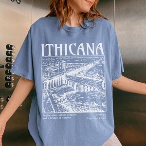 May include: A light blue t-shirt with a white graphic print of a bridge and the text "ITHICANA". The text below the graphic reads "Raging seas, hidden jungles and a bridge of secrets.  Roled by King fun Lestell."