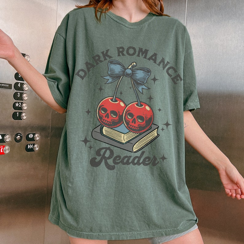 K&ouml;nnte beinhalten: Gr&uuml;nes T-Shirt mit dem Text "DARK ROMANCE Reader". Die Grafik zeigt zwei rote Kirschen mit Totenkopfgesichtern, eine blaue Schleife und einen Stapel B&uuml;cher. Das Design enth&auml;lt Sterne.