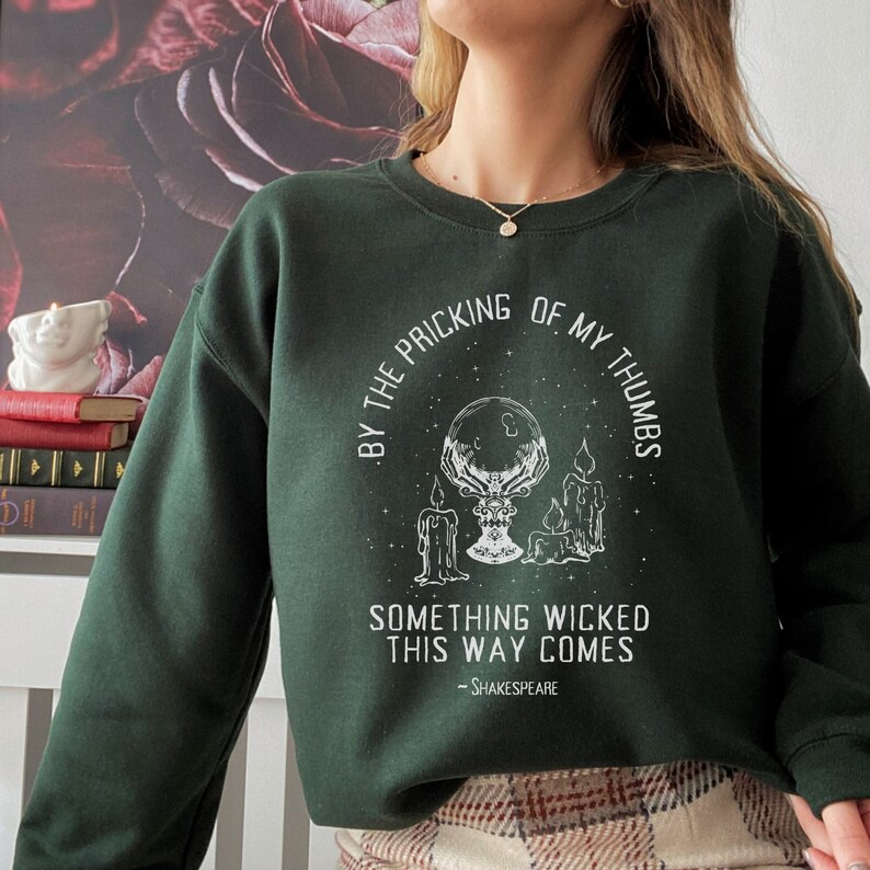 Op de afbeelding: Een donkergroen sweatshirt met een witte grafische print van een kristallen bol, kaarsen en de tekst "By the pricking of my thumbs, something wicked this way comes - Shakespeare".
