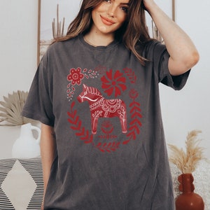 May include: A grey t-shirt featuring a red and white Scandinavian Dala horse design. The horse is surrounded by red floral patterns.