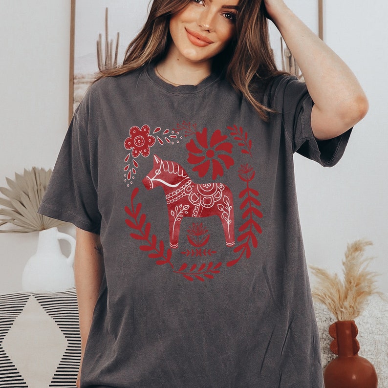 May include: A grey t-shirt featuring a red and white Scandinavian Dala horse design. The horse is surrounded by red floral patterns.
