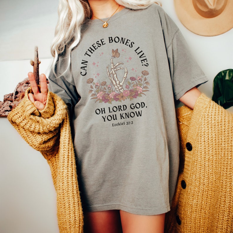 Puede incluir: Camiseta gris con una mano de esqueleto sosteniendo una mariposa y detalles florales. El texto dice "Can these bones live? Oh Lord God, you know. Ezekiel 37:3"