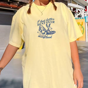 K&ouml;nnte beinhalten: Hellgelbes T-Shirt mit marineblauem Aufdruck. Der Aufdruck lautet "I look better bent over a book" und zeigt eine Vintage-Illustration einer lesenden Frau. Das T-Shirt hat kurze &Auml;rmel und eine lockere Passform.