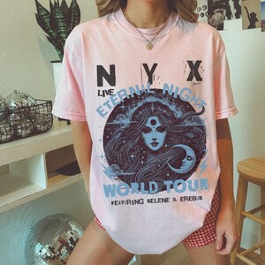 May include: A pale pink t-shirt with the text "NYX LIVE ETERNAL NIGHT WORLD TOUR" in black and light blue. The graphic features a woman's face with long hair, a crescent moon, and stars. The shirt is short-sleeved.