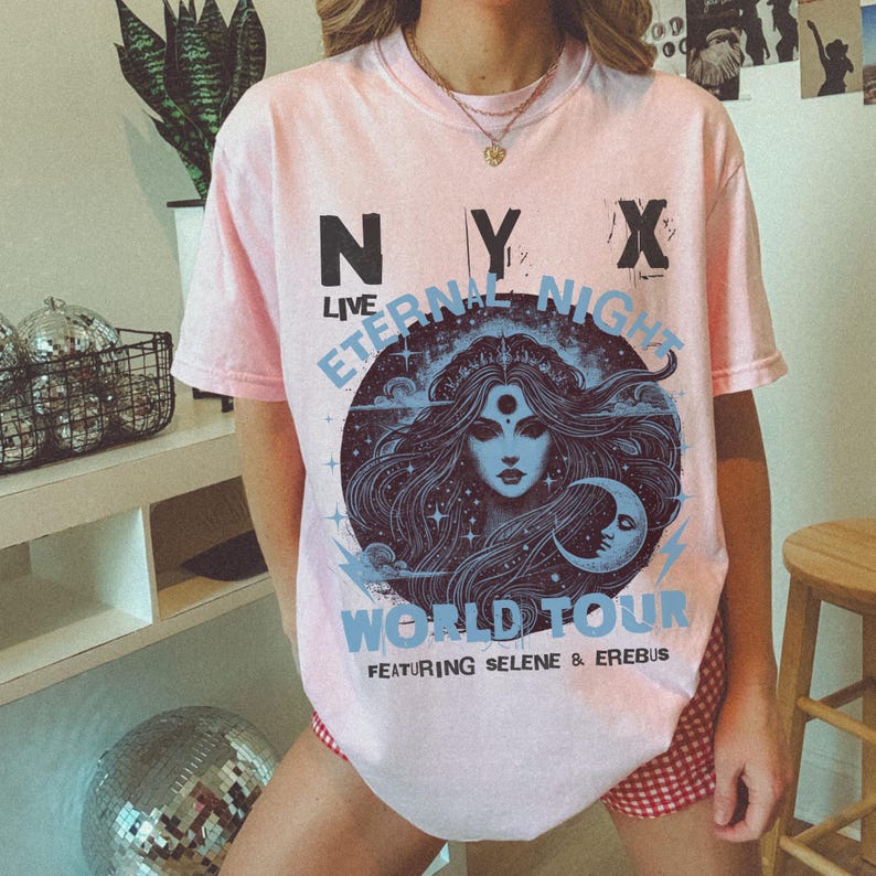 May include: A pale pink t-shirt with the text "NYX LIVE ETERNAL NIGHT WORLD TOUR" in black and light blue. The graphic features a woman's face with long hair, a crescent moon, and stars. The shirt is short-sleeved.