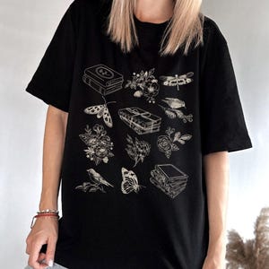 May include: Black t-shirt with a white graphic design featuring a stack of books, flowers, a butterfly, a dragonfly, and a bird.