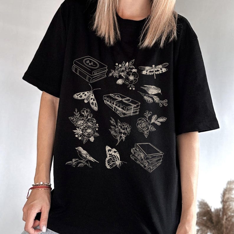 May include: Black t-shirt with a white graphic design featuring a stack of books, flowers, a butterfly, a dragonfly, and a bird.