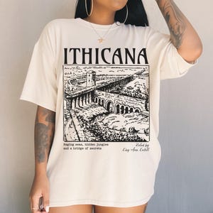 May include: A white t-shirt with a black graphic print of a bridge and a castle. The text "ITHICANA" is printed above the graphic. The text "Raging seas, hidden jungles and a bridge of secrets" is printed below the graphic. The text "Roled by King Aren Keitell" is printed in the bottom right corner.