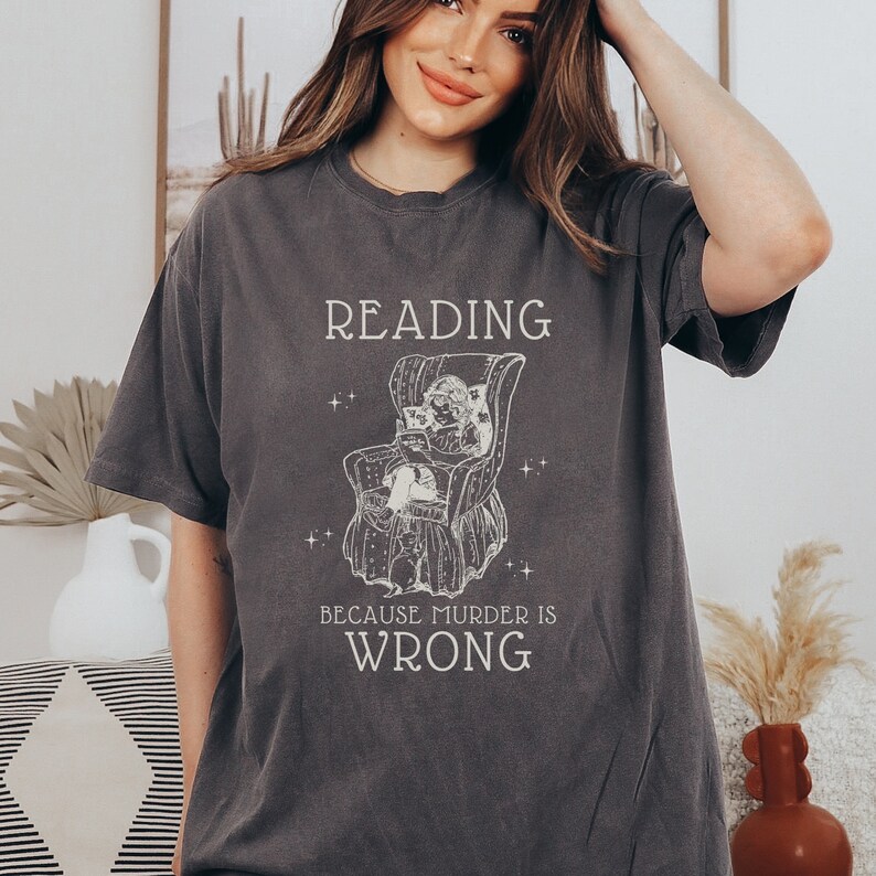Puede incluir: Una camiseta gris oscuro con un gr&aacute;fico blanco de una persona leyendo en una silla. El texto en la camiseta dice "Reading because murder is wrong."