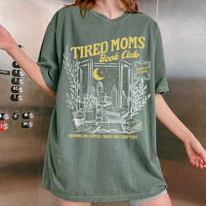 May include: Green t-shirt with a white graphic of a window with a cityscape, moon, and stars. The text "TIRED MOMS Book Club" is in yellow above the window. The text "RUNNING ON COFFEE, CHAOS AND CHAPTERS" is in yellow below the window. The text "NIGHTLY AT 8PM" is in yellow in the upper right corner of the window.