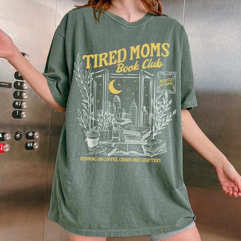 May include: Green t-shirt with a white graphic of a window with a cityscape, moon, and stars. The text "TIRED MOMS Book Club" is in yellow above the window. The text "RUNNING ON COFFEE, CHAOS AND CHAPTERS" is in yellow below the window. The text "NIGHTLY AT 8PM" is in yellow in the upper right corner of the window.