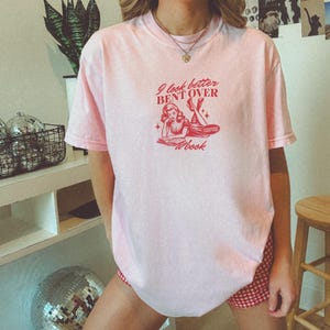 K&ouml;nnte beinhalten: Hellrosa T-Shirt mit rotem Aufdruck "I look better BENT OVER a book". Die Grafik zeigt eine Frau beim Lesen. Das Shirt hat kurze &Auml;rmel und einen Rundhalsausschnitt.