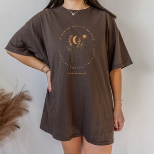 May include: A brown oversized t-shirt with a graphic of two hands holding a crescent moon and sun with a quote from Emily Dickinson that reads "I took my power in my hand and went against the world".