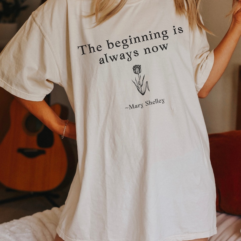 May include: White oversized t-shirt with the text "The beginning is always now" and a small flower graphic. The text "~Mary Shelley" is below the graphic.