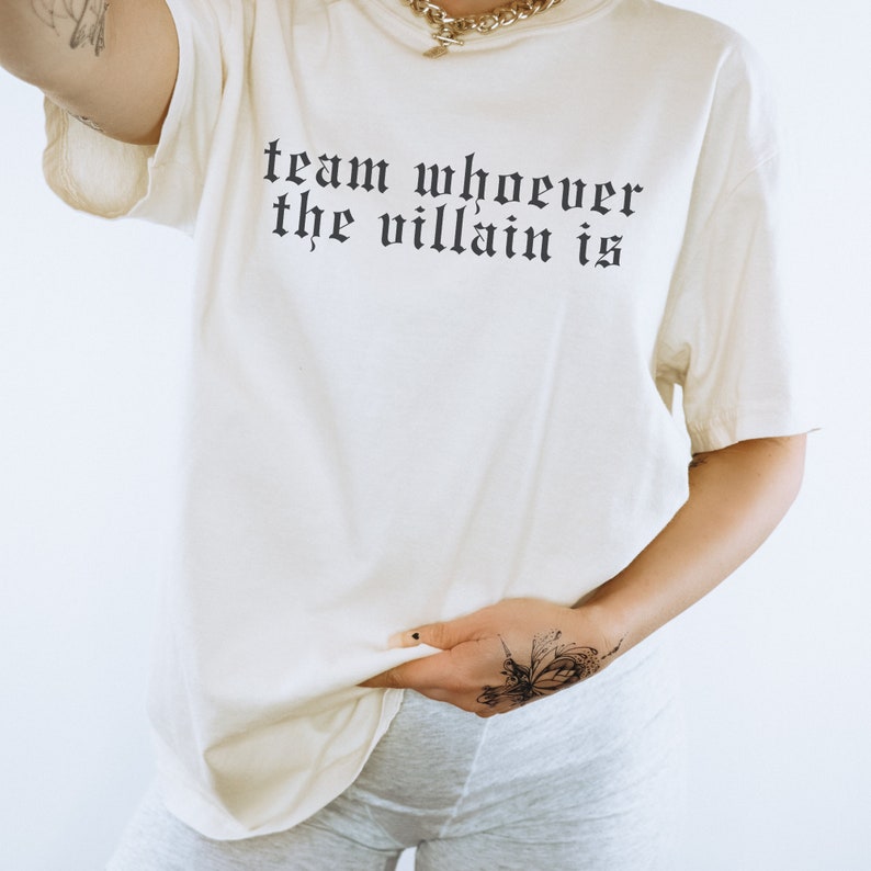 May include: A white t-shirt with the text "team whoever the villain is" printed in black. The shirt is being worn by a person with a butterfly tattoo on their wrist.
