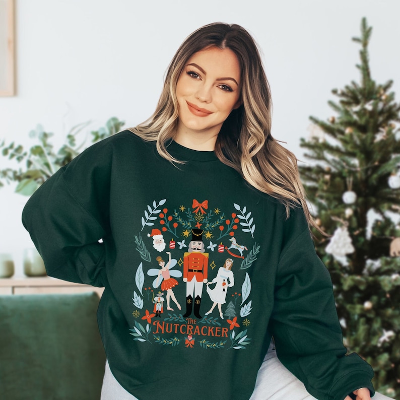 May include: A dark green sweatshirt with a festive design featuring a Nutcracker, a ballerina, and other characters. The design is surrounded by a wreath of greenery and snowflakes. The text "The Nutcracker" is printed below the design.