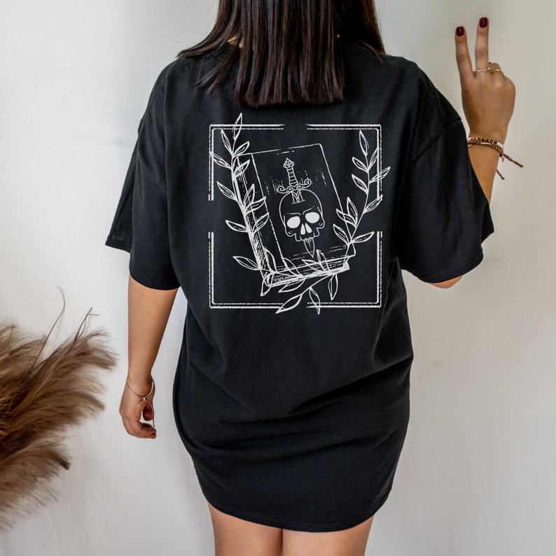 May include: Black oversized t-shirt with a white graphic of a book with a skull and a dagger inside. The book is surrounded by leaves.