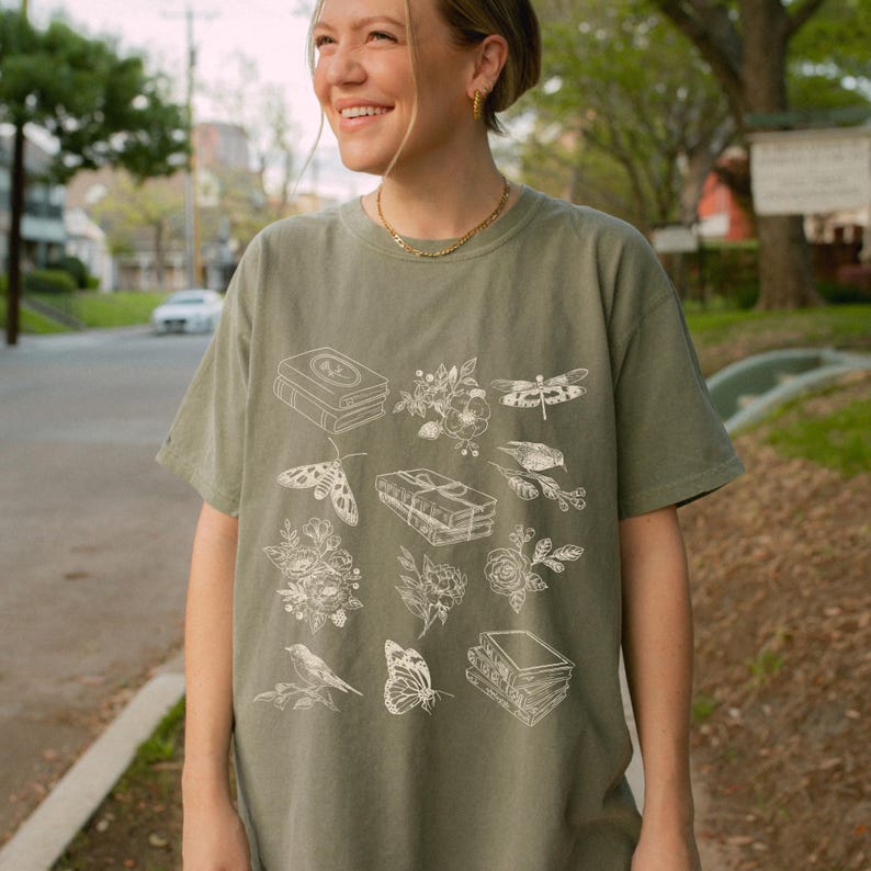 May include: A sage green t-shirt with a white graphic print of books, flowers, butterflies, and dragonflies.