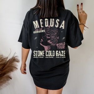 May include: Black t-shirt with a graphic print of Medusa, a mythical creature with snakes for hair. The text "Medusa and the Gorgons World Tour Stone Cold Gaze" is printed below the image. The text "Serpent's Scream Hades Inferno Stare of Justice" is printed below the graphic.
