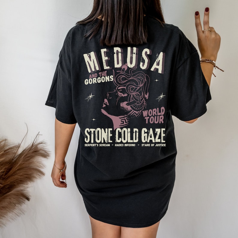 May include: Black t-shirt with a graphic print of Medusa, a mythical creature with snakes for hair. The text "Medusa and the Gorgons World Tour Stone Cold Gaze" is printed below the image. The text "Serpent's Scream Hades Inferno Stare of Justice" is printed below the graphic.