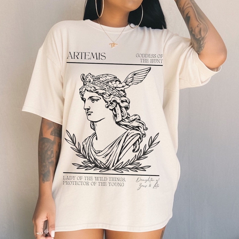 May include: A white oversized t-shirt with a black graphic print of Artemis, the Greek goddess of the hunt. The graphic includes a drawing of Artemis with a crown and wings, surrounded by laurel leaves. The text on the shirt reads "ARTEMIS, GODDESS OF THE HUNT, LADY OF THE WILD THINGS, PROTECTOR OF THE YOUNG, Daughter of Zeus & Leto."