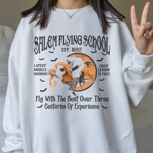 Op de afbeelding: Wit sweatshirt met een zwarte en oranje grafische print van een heks die op een bezem over een halve maan vliegt. De tekst luidt "Salem Flying School Est. 1692" en "Fly With The Best Over Three Centuries Of Experience".