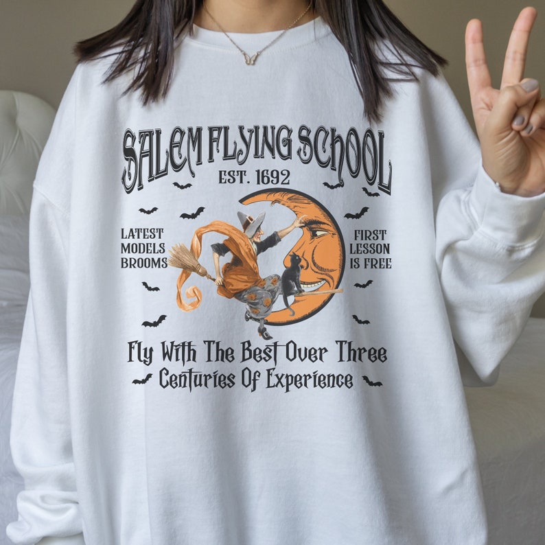 Op de afbeelding: Wit sweatshirt met een zwarte en oranje grafische print van een heks die op een bezem over een halve maan vliegt. De tekst luidt "Salem Flying School Est. 1692" en "Fly With The Best Over Three Centuries Of Experience".