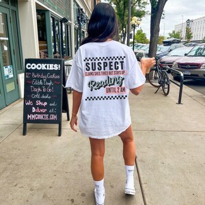 May include: White t-shirt with black and white checkered trim and the text "SUSPECT CLAIMS SHES TIRED BUT STAYS UP Reading UNTIL 2 AM" in black and pink lettering.