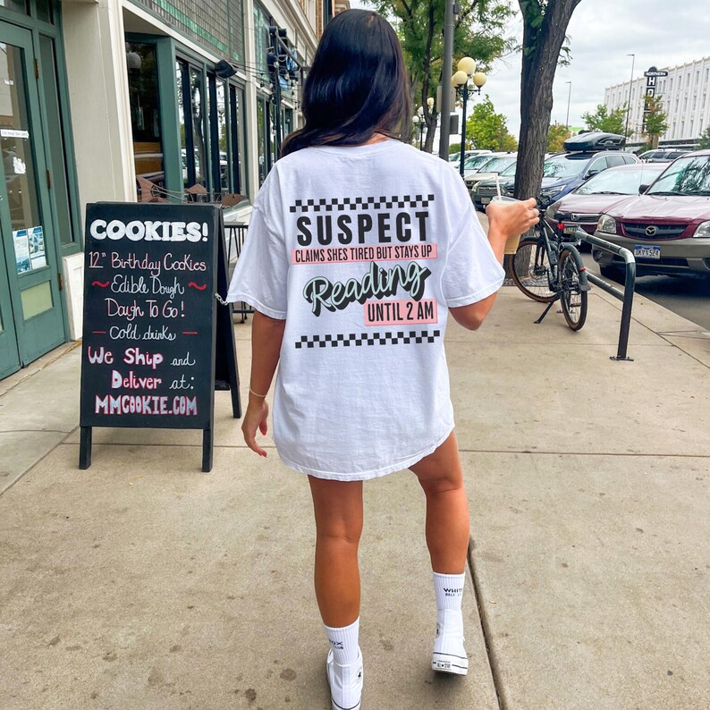 May include: White t-shirt with black and white checkered trim and the text "SUSPECT CLAIMS SHES TIRED BUT STAYS UP Reading UNTIL 2 AM" in black and pink lettering.