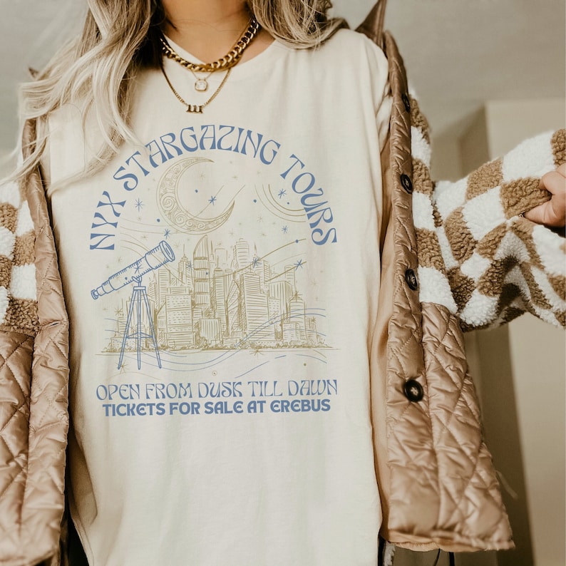 K&ouml;nnte beinhalten: Ein wei&szlig;es T-Shirt mit einem blauen Grafikdesign eines Stadtbildes mit einer zunehmenden Mondsichel und Sternen. Der Text "NYX STARGAZING TOURS" ist in einem Kreis um den Mond geschrieben. Der Text "OPEN FROM DUSK TILL DAWN TICKETS FOR SALE AT EREBUS" steht unter der Grafik.