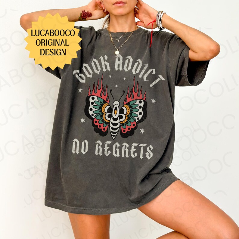 May include: Dark gray t-shirt with the text "BOOK ADDICT NO REGRETS" and a butterfly graphic. The butterfly has teal, red, and yellow details with flames. The shirt also has the text "LUCABOOCO ORIGINAL DESIGN".