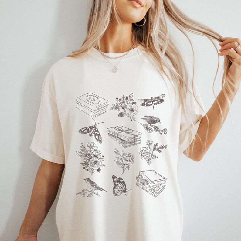 May include: A white t-shirt with a black and white graphic design featuring books, flowers, butterflies, and dragonflies.